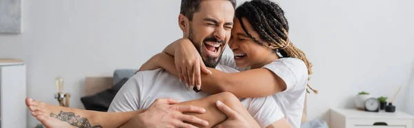Überglückliches gemischtrassiges Paar in weißen T-Shirts, das sich umarmt und mit geschlossenen Augen im heimischen Schlafzimmer lacht, Banner — Stockfoto