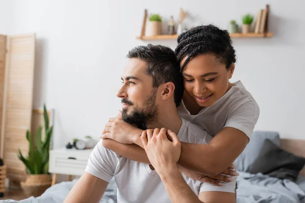 Happy african american woman embracing dreamy bearded man looking away in bedroom at home — Stock Photo