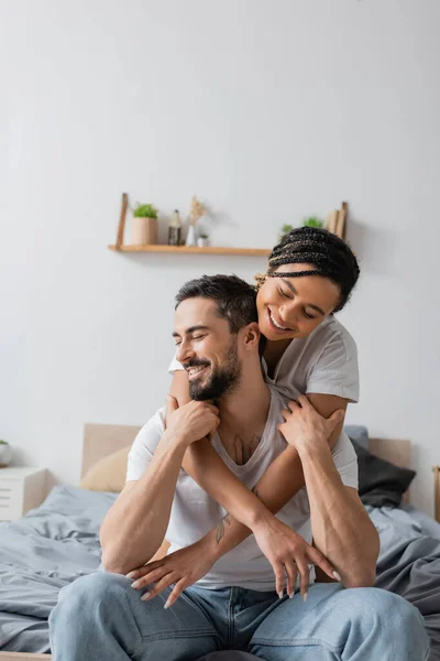 Pleased african american woman hugging bearded man smiling with closed eyes in bedroom at home — Stock Photo