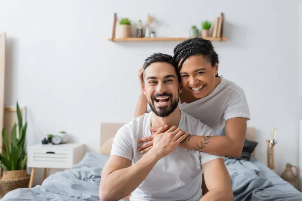 Super alegre casal multiétnico em t-shirts brancas rindo da câmera no quarto em casa — Fotografia de Stock