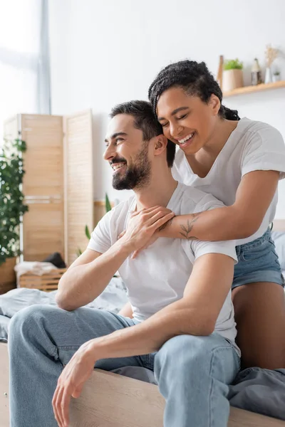 Mulher americana africana alegre com olhos fechados abraçando namorado feliz em t-shirt branca e jeans sentados na cama em casa — Fotografia de Stock