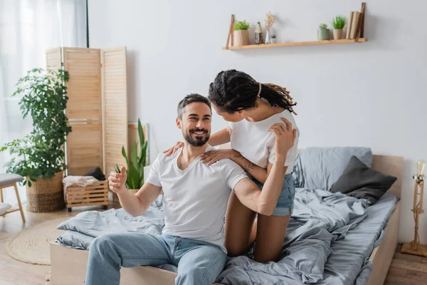 Young african american woman embracing carefree bearded man sitting in white t-shirt and jeans in modern bedroom — Stock Photo