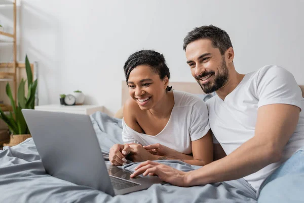 Joyful interracial couple in white t-shirts watching movie on laptop in bedroom at home — Stock Photo