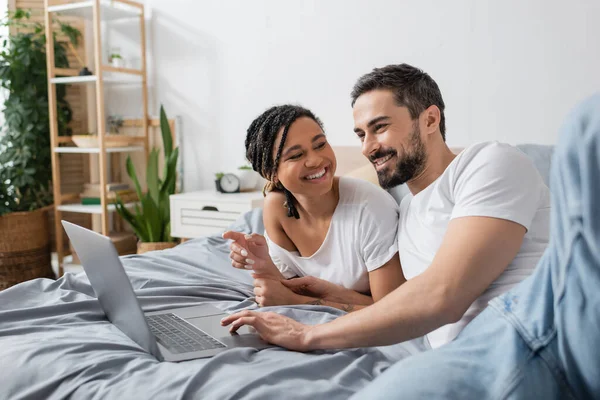 Allegra donna afro-americana che indica il computer portatile e guarda l'uomo barbuto sorridente sul letto a casa — Foto stock