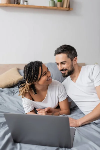 Feliz pareja multiétnica en camisetas blancas sonriendo el uno al otro cerca de la computadora portátil en la cama en casa - foto de stock