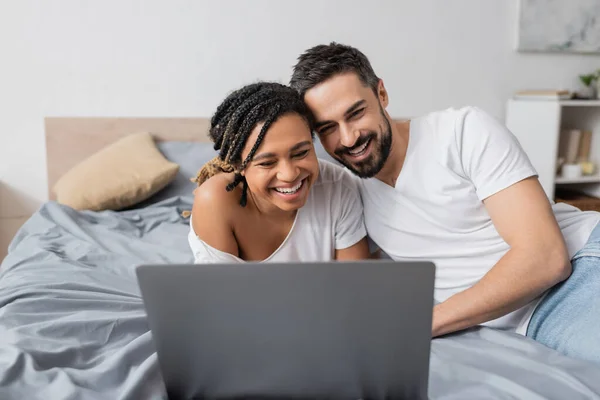 Возбужденная межрасовая пара смеется возле ноутбука во время просмотра фильма на кровати дома — стоковое фото