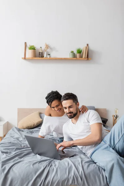 Femme afro-américaine souriante embrassant homme barbu tout en regardant un film sur ordinateur portable dans la chambre à coucher à la maison — Photo de stock