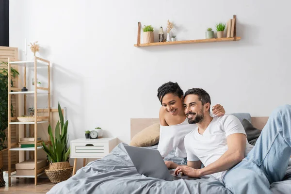 Беззаботная межрасовая пара улыбается возле ноутбука во время просмотра фильма в современной спальне дома — стоковое фото