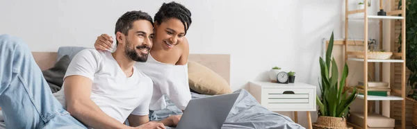Jeune femme afro-américaine et homme barbu en t-shirts blancs regardant un film sur ordinateur portable dans la chambre, bannière — Photo de stock