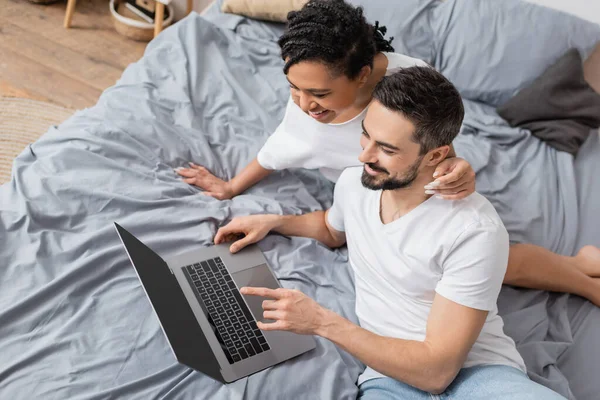 Vista ad alto angolo di uomo barbuto allegro e donna afroamericana guardando film sul computer sul letto a casa — Foto stock