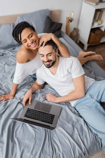 Высокий угол обзора веселой межрасовой пары в белых футболках, смотрящей фильм на ноутбуке на кровати дома — стоковое фото