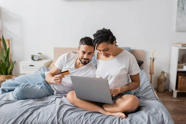Homem barbudo segurando cartão de crédito perto de mulher americana africana sentado com pernas cruzadas e usando laptop na cama em casa — Fotografia de Stock
