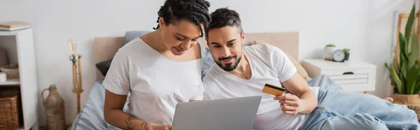 Smiling bearded man holding credit card near african american woman with laptop in bedroom at home, banner — Stock Photo