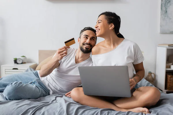 Hombre barbudo feliz sosteniendo la tarjeta de crédito cerca de la novia afroamericana riendo sentado con el ordenador portátil en la cama en casa - foto de stock