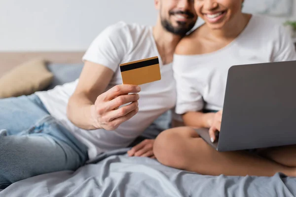 Обрезанный вид размытого мужчины, держащего кредитную карту рядом с улыбающейся африканской американкой с ноутбуком в спальне дома — стоковое фото