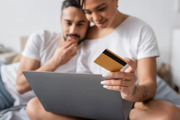 Enfoque selectivo de la tarjeta de crédito y el ordenador portátil cerca borrosa pareja interracial pensando durante las compras en línea en el dormitorio en casa - foto de stock