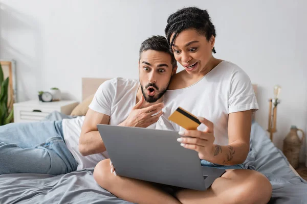 Изумленная африканская американка с ноутбуком и кредитной картой рядом с мужчиной с открытым ртом на кровати дома — стоковое фото