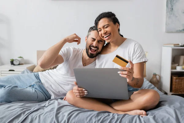 Overjoyed interracial couple laughing with closed eyes near laptop and credit card on bed at home — Stock Photo