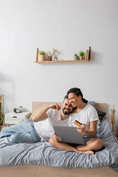 Allegra coppia multietnica con computer portatile e carta di credito ridere durante lo shopping online sul letto a casa — Foto stock