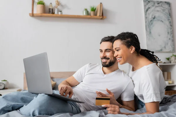 Cheerful bearded man using laptop near african american woman with credit card on bed at home — Stock Photo