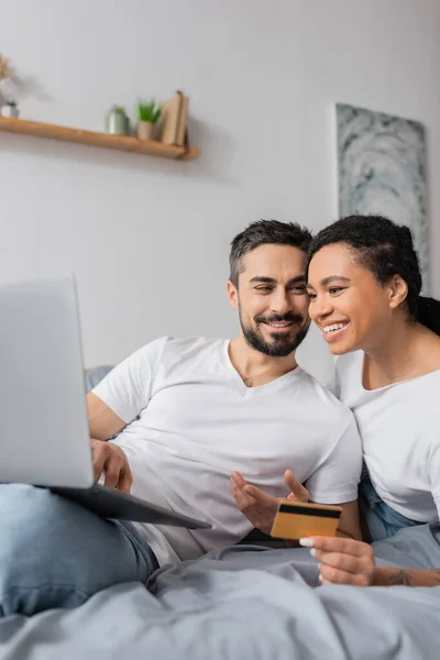 Homem despreocupado apontando para laptop perto de mulher afro-americana com cartão de crédito durante compras on-line no quarto em casa — Fotografia de Stock