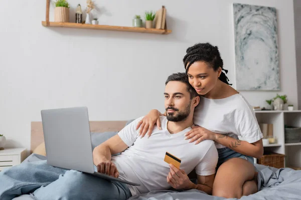 Young african american woman hugging shoulders of bearded man with laptop and credit card on bed at home — Stock Photo