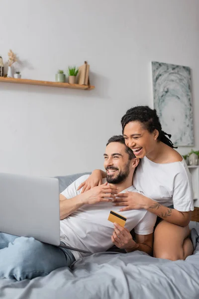 Eccitata coppia multietnica con laptop e carta di credito ridere durante lo shopping online in camera da letto a casa — Foto stock