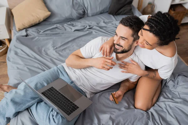 Visão de alto ângulo da mulher americana africana alegre abraçando homem barbudo com laptop e cartão de crédito na cama em casa — Fotografia de Stock