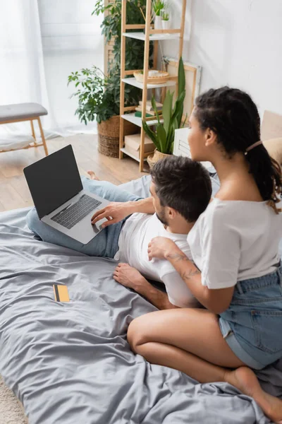 Interracial couple looking at laptop with blank screen near credit card on bed at home — Stock Photo
