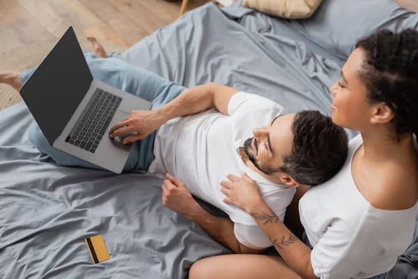 Vue du haut de sourire homme barbu en utilisant un ordinateur portable près de petite amie afro-américaine et carte de crédit sur le lit à la maison — Photo de stock