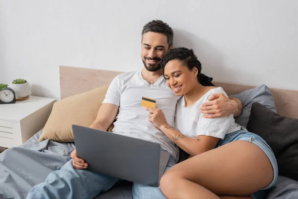 Alegre barbudo hombre con portátil abrazando afroamericano novia celebración de tarjeta de crédito en la cama en casa - foto de stock