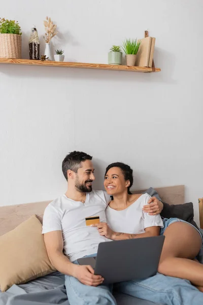 Feliz pareja interracial con portátil y tarjeta de crédito mirándose en el dormitorio en casa - foto de stock