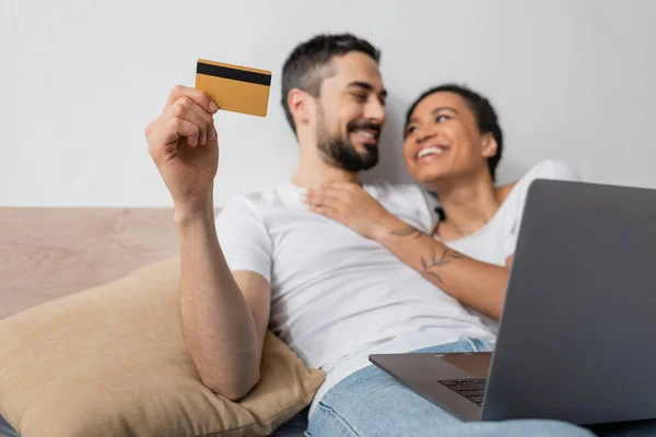 Hombre barbudo alegre celebración de la tarjeta de crédito cerca de la computadora portátil y la mujer afroamericana feliz en el dormitorio en casa - foto de stock