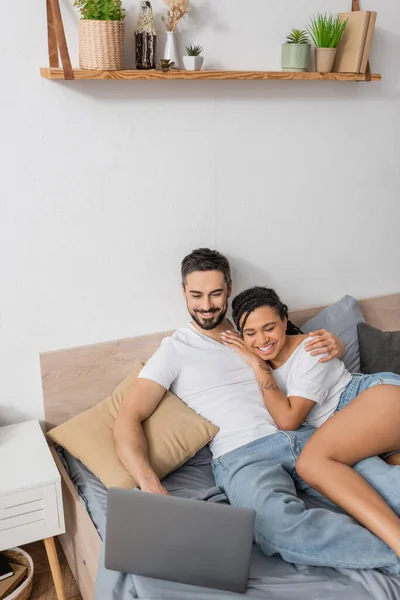 Vue grand angle de joyeux couple interracial en t-shirts blancs regarder film sur ordinateur portable sur le lit à la maison — Photo de stock
