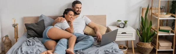 Весела афроамериканська жінка з бородатим хлопцем дивиться фільм на ноутбуці в сучасній спальні вдома, банер — стокове фото