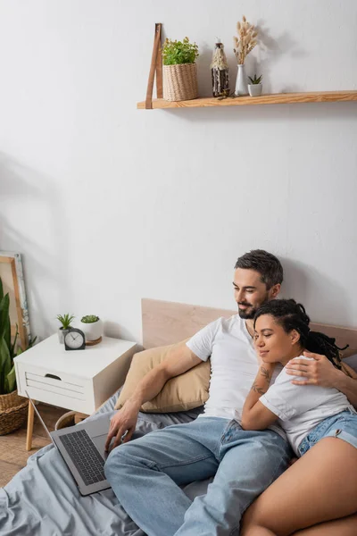 Pleased bearded man and young african american woman watching movie on laptop on bed at home — Stock Photo