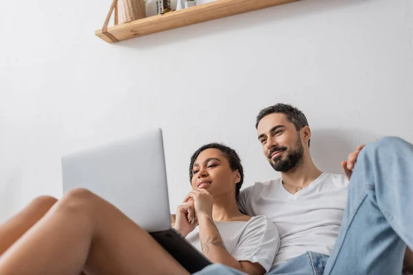 Baixo ângulo de visão de casal multiétnico sorridente assistindo filme no laptop enquanto descansa no quarto em casa — Fotografia de Stock