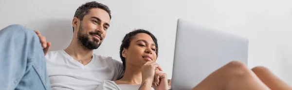 Low angle view of interracial couple smiling while watching movie on laptop at home, banner — Stock Photo