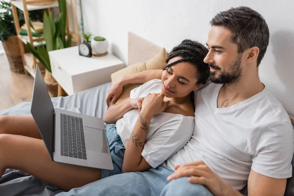 Беззаботная межрасовая пара в белых футболках смотрит кино на ноутбуке в спальне дома — стоковое фото