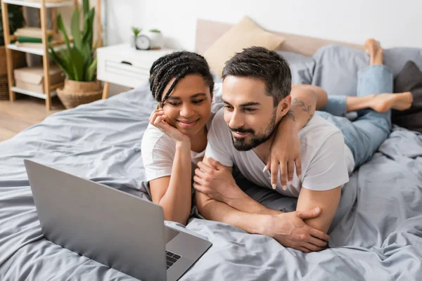 Heureuse femme afro-américaine étreignant homme barbu tout en regardant un film sur ordinateur portable dans la chambre à coucher à la maison — Photo de stock