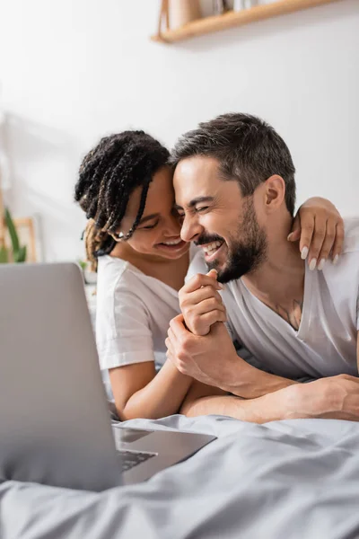 Aufgeregtes interrassisches Paar lacht mit geschlossenen Augen, während es im Schlafzimmer einen Komödienfilm auf dem Laptop anschaut — Stockfoto