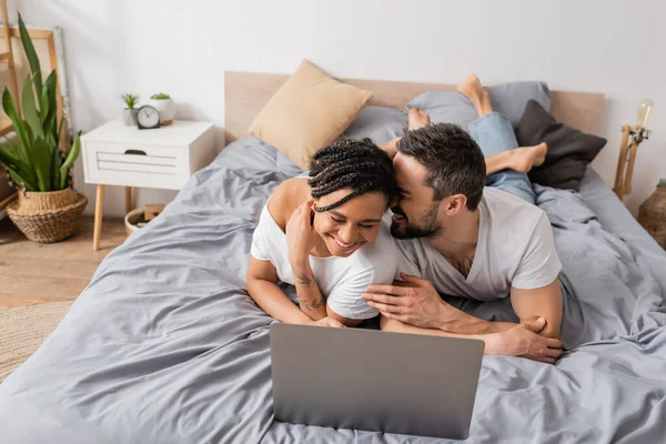 Overjoyed interracial couple laughing with closed eyes while watching film on laptop in bedroom at home — Stock Photo