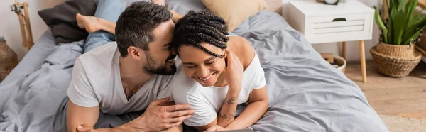Alegre hombre diciendo secreto a la joven afroamericana mujer mientras se relaja en la cama en casa, pancarta - foto de stock