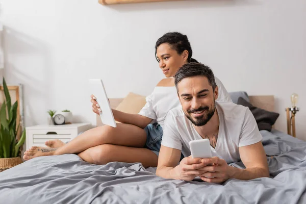 Carefree interracial couple in white t-shirts using gadgets while resting on bed at home — Stock Photo