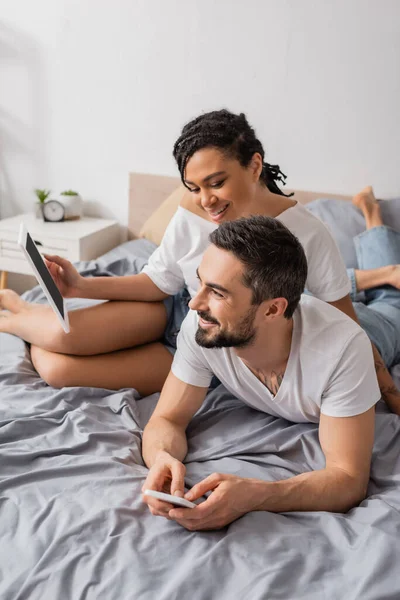 Joyeuse femme afro-américaine tenant tablette numérique près de sourire homme barbu couché avec smartphone sur le lit à la maison — Photo de stock