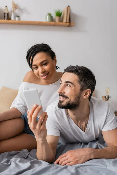 Cheerful bearded man showing smartphone to smiling african american woman while resting on bed at home — Stock Photo