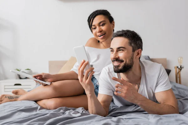 Веселий бородатий чоловік вказує на мобільний телефон біля здивованої афроамериканської жінки, що сидить з цифровим планшетом на ліжку — стокове фото