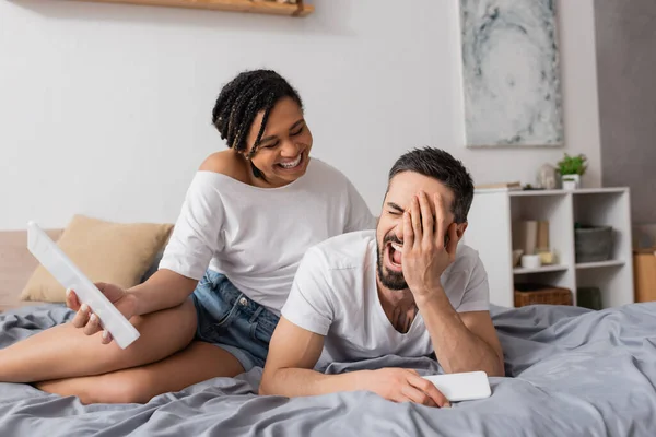 Hombre excitado cubriendo la cara y riendo cerca de la novia afroamericana con tableta digital en la cama en casa - foto de stock