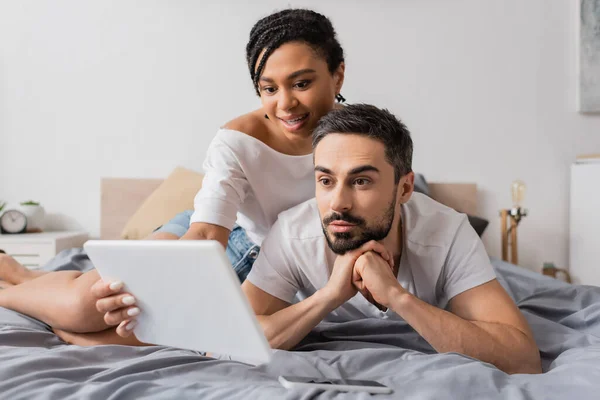 Beeindruckte Afroamerikanerin zeigt überraschtem bärtigen Mann, der zu Hause im Bett liegt, digitales Tablet — Stockfoto