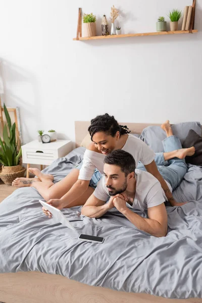 Улыбающаяся африканская американка, показывающая цифровой планшет изумленному мужчине, отдыхающему дома на кровати — стоковое фото